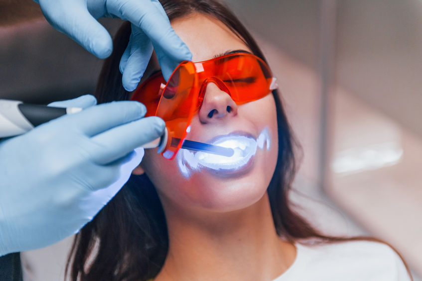 A person undergoing a dental procedure with protective eyewear and a dental tool emitting blue light in their mouth.